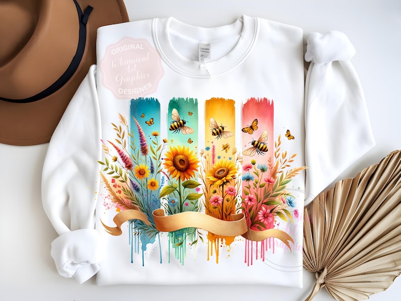 May include: White sweatshirt featuring a colorful floral and bee design. The design includes sunflowers, wildflowers, and butterflies, with a gold ribbon across the bottom. The words "Original Whimsical Art Graphics Designer" are on the upper left.