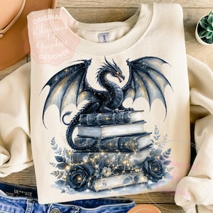 Könnte beinhalten: Cremefarbenes Sweatshirt mit einem schwarzen Drachen mit ausgebreiteten Flügeln, der auf einem Stapel Bücher sitzt. Das Design umfasst blaue Rosen, florale Akzente und den Text "ORIGINAL Whimsical Art Graphic DESIGNER".
