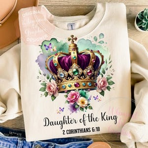 Op de afbeelding: Crèmekleurige sweatshirt met een koninklijk kroonontwerp met paarse, gouden en juwelen accenten, omringd door roze en paarse bloemen en vlinders. De tekst "Daughter of the King" en "2 Corinthians 6:18" is eronder gedrukt.