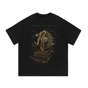 Puede incluir: Una camiseta negra de manga corta con un gráfico dorado y plateado en el pecho. El diseño incluye el texto "DIAMONDS ARE FOREVER" sobre un logotipo "R" estilizado y un emblema circular, con el texto "ROC-A-FELLA RECORDS" impreso debajo.