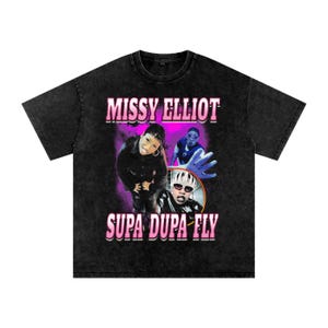 May include: Black t-shirt featuring a graphic of Missy Elliott with the text "MISSY ELLIOT" above and "SUPA DUPA FLY" below. The design includes images of Missy Elliott and a purple and pink color scheme.