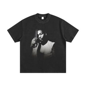 Può includere: T-shirt nero con immagine in bianco e nero di una persona che tiene una chitarra. La persona indossa una canotta bianca. La t-shirt ha girocollo e maniche corte.