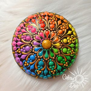 Peut inclure: Une pierre ronde noire ornée d'un motif mandala vibrant peint avec des points en relief dans un spectre arc-en-ciel de couleurs incluant rouge, orange, jaune, vert, bleu et violet, rehaussé de détails dorés et d'un point central doré.