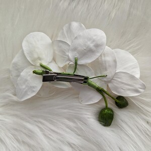 Op de afbeelding: Een witte orchidee haarspeld met een groene steel en bladeren. De speld is gemaakt van metaal en heeft een veermechanisme.