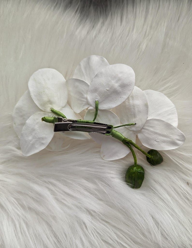 Op de afbeelding: Een witte orchidee haarspeld met een groene steel en bladeren. De speld is gemaakt van metaal en heeft een veermechanisme.