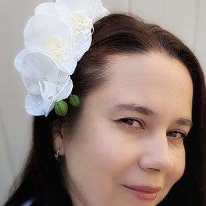 Op de afbeelding: Witte orchidee haarspeld met drie bloemen en groene bladeren. De speld wordt in het haar van een vrouw gedragen.