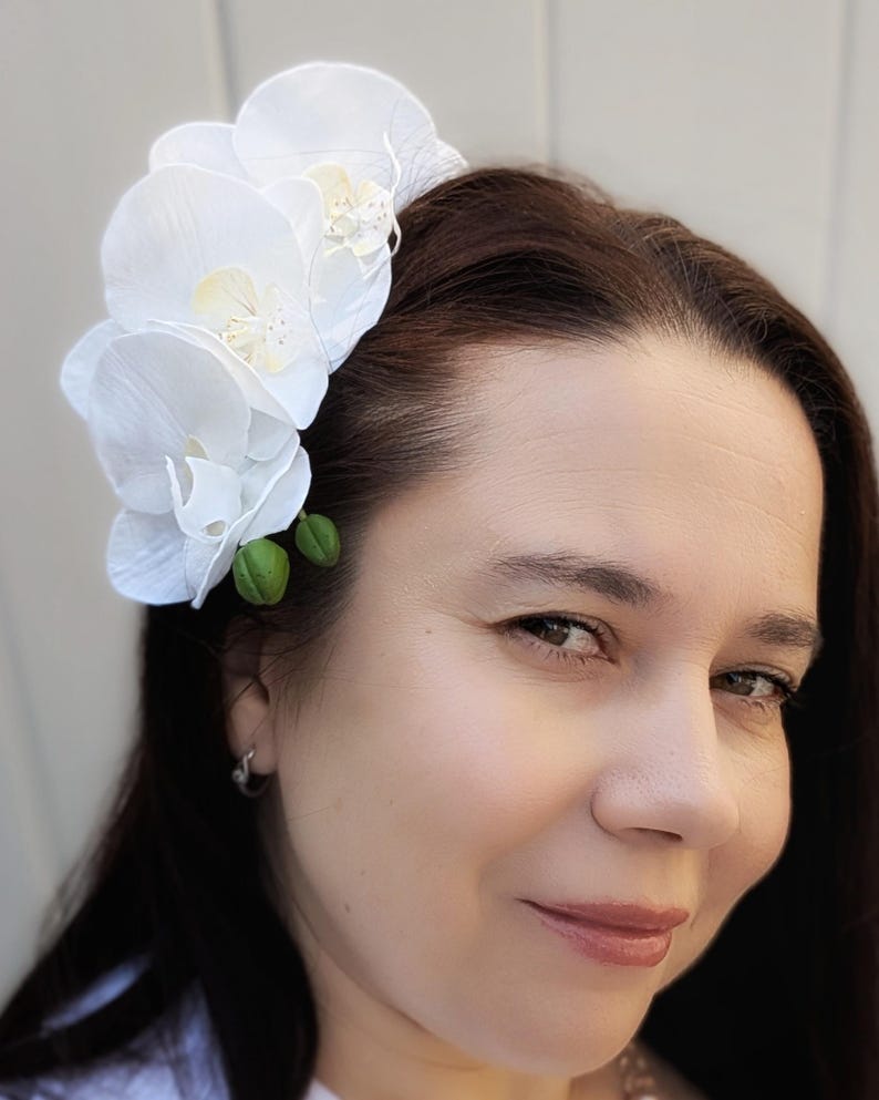 Op de afbeelding: Witte orchidee haarspeld met drie bloemen en groene bladeren. De speld wordt in het haar van een vrouw gedragen.