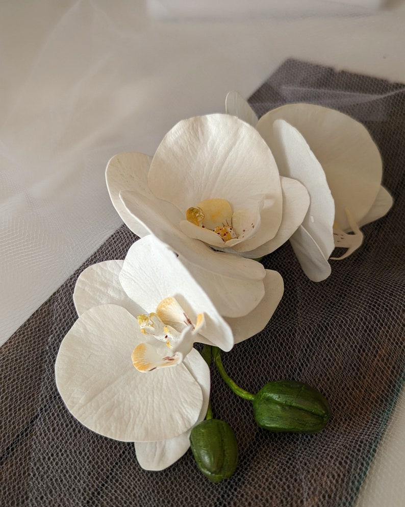 Op de afbeelding: Drie witte orchidee&euml;n met groene stelen en knoppen op een grijze, textuurrijke achtergrond.