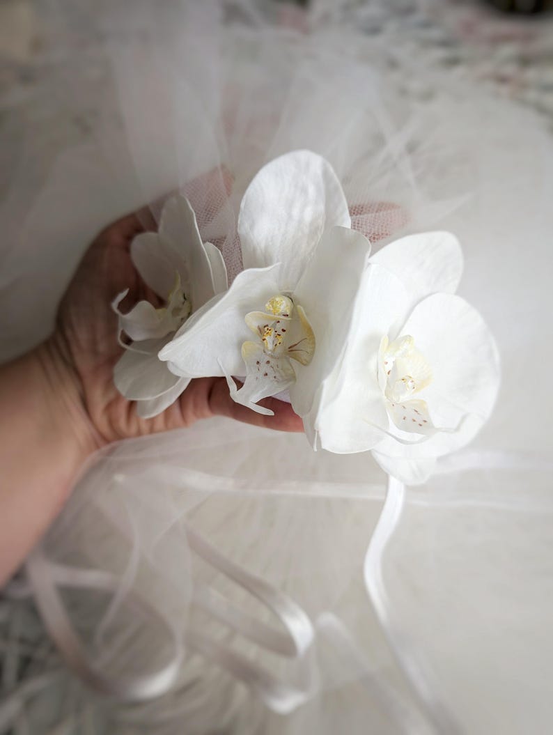 Op de afbeelding: Drie witte orchidee&euml;n met gele centra worden in een hand gehouden. De bloemen bevinden zich op een witte tule achtergrond.
