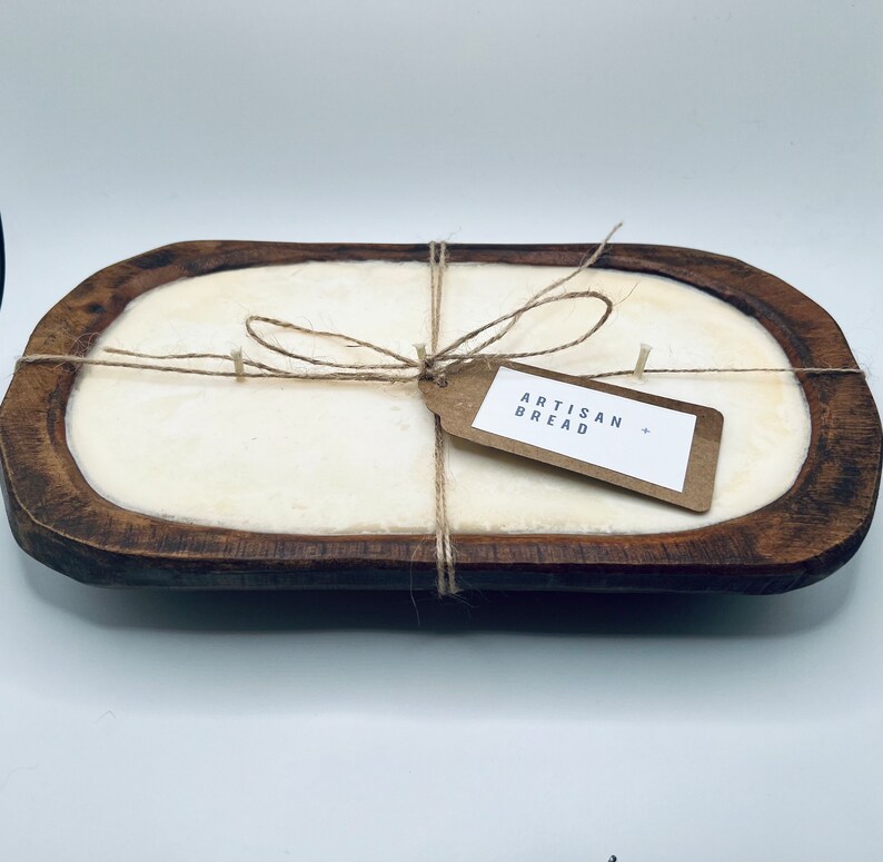 May include: A wooden bread bowl with a white candle inside. The candle is tied with twine and a brown paper tag that says "Artisan Bread".