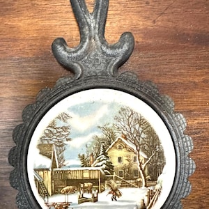 Puede incluir: Un salvamanteles decorativo con una escena invernal. El inserto cerámico redondo presenta una granja cubierta de nieve con una casa, un granero y figuras. El salvamanteles tiene un borde festoneado y un asa, todo en un acabado oscuro. El texto "THE FARMER'S HOME-WINTER" es visible.