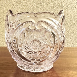 Puede incluir: Un cuenco de vidrio transparente y decorativo con intrincados patrones grabados. El cuenco presenta un borde festoneado y un diseño detallado de líneas arremolinadas y formas de estrella. El vidrio tiene un ligero tono rosado y el cuenco se asienta sobre una pequeña base redonda.