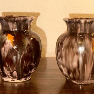 Puede incluir: Dos pequeños jarrones de cerámica a juego con un acabado brillante y veteado. Los jarrones son principalmente de color marrón oscuro con vetas blancas y naranjas. Tienen un cuerpo redondeado y un borde acampanado, adecuados para exhibir flores pequeñas o artículos decorativos.