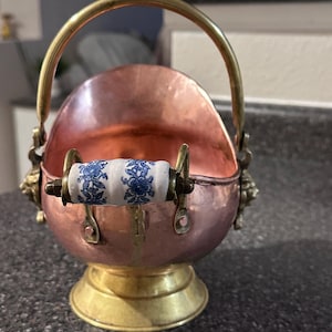 May include: A copper-coloured metal coal scuttle with a brass handle and base. The handle has a white ceramic grip with blue floral designs. The scuttle has decorative lion head accents on each side.
