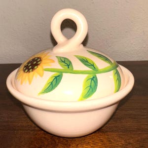 May include: A light pink ceramic bowl with a matching lid. The lid has a yellow sunflower design with green leaves and a loop handle. The bowl is likely for serving or storage.