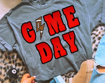 Op de afbeelding: Grijze Game Day t-shirt met rode letters en een voetbal-bliksem graphic. Een beige hoed met de tekst "Brunettes Have More Fun" is zichtbaar. De afbeelding bevat ook een blauwe jeans en een discobal.