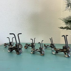 Puede incluir: Una colección de pequeñas esculturas de motocicletas de metal hechas a mano. Cada moto en miniatura presenta un diseño único, con manillares, asientos y ruedas construidos con tuercas, pernos y otros herrajes. Las esculturas están dispuestas sobre una superficie azul claro.