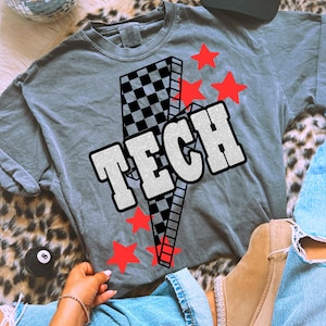 May include: Gray t-shirt with a graphic design featuring the word "TECH" in a bold, outlined font, a lightning bolt, red stars, and a checkered pattern. A hat that says "Brunettes Have More Fun" is also visible.