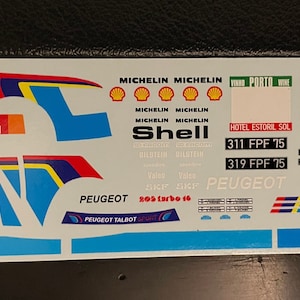 Peut inclure: Planche de décalcomanies pour voiture de course Peugeot. Les décalcomanies présentent les logos Michelin et Shell, ainsi que divers graphismes de course en bleu, rouge, jaune et blanc. Le texte comprend "PEUGEOT", "TALBOT" et des numéros.