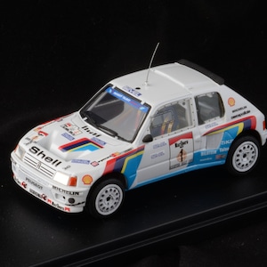 Peut inclure: Modèle réduit de voiture de rallye Peugeot blanche, ornée d'autocollants de sponsors tels que Shell, Marlboro et Bilstein. Le modèle présente des accents bleus, rouges et jaunes, une antenne de toit et des roues blanches. La voiture porte le numéro 1.
