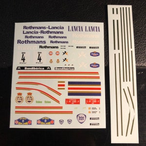 Puede incluir: Una hoja de calcomanías blancas con varios logotipos y texto, incluyendo "Rothmans-Lancia", "Lancia-Rothmans", "Rothmans", "Bendibérica", "Valeo", "Pirelli", "Gulf", y "Lancia".