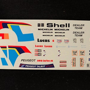 May include: A white decal sheet with various stickers for a model car. The stickers include the words "Shell", "Michelin", "Lucas", "Peugeot Talbot", and "B555 SRW". There are also numbers "7" and "55" on the sheet.