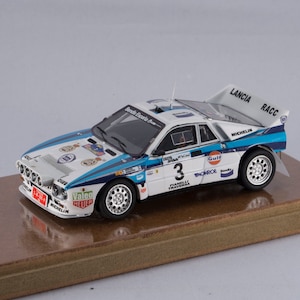 May include: A 1/18 scale diecast model of a Lancia 037 rally car, painted white, blue and red. The car has the number "3" on the side and sponsors such as "Gulf", "Valeo", "Michelin", and "Bendix" on the body. The car is displayed on a wooden base.