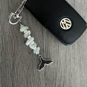 Op de afbeelding: Zwarte autosleutel met een zilveren VW-logo en een decoratieve sleutelhanger. De sleutelhanger heeft een streng van lichtblauwe en witte stenen en een zilveren walvisstaartbedel.
