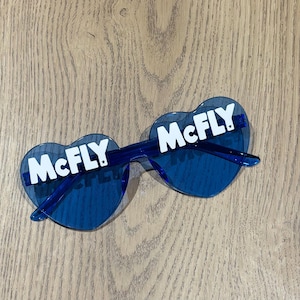 May include: Blue heart-shaped sunglasses with the word "McFLY" in white across each lens. The sunglasses have a transparent blue frame and are set against a light wood grain background.