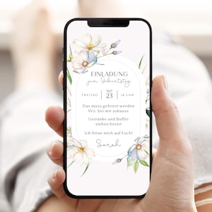 May include: A smartphone screen shows a digital invitation with floral decorations. The design features white flowers with blue and green watercolour details. Text includes "EINLADUNG zum Geburtstag", date, time, and location details. The invitation is held in a hand.