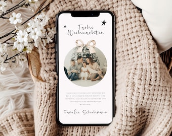 以下が含まれることがあります： スマートフォンには「Frohe Weihnachten」の文字と家族写真が入ったクリスマスカードが表示されています。カードはベージュのニットブランケットの上に置かれ、背景には白い花があります。