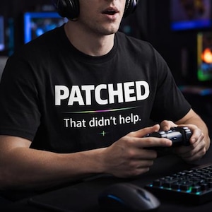 May include: Black t-shirt with the word "PATCHED" in large white letters. Below, the text "That didn't help." is printed in white, with a colourful line above it. A person is wearing headphones and holding a game controller.