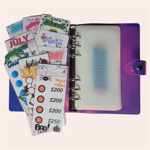 May include: A purple binder with a clear pocket labeled "January" and several monthly dividers. The dividers are labeled with months like "July," "March," and "October," and include decorative elements and text. The binder is designed for budgeting.