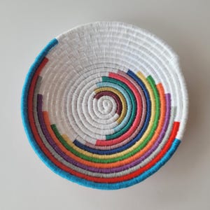 May include: A round, shallow bowl constructed from coiled white rope. The interior features a spiral pattern of multicolored thread accents in shades of blue, green, yellow, orange, red, and purple, creating a vibrant, concentric circular design.