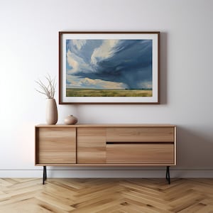 May include: A framed painting of a stormy sky over a grassy field. The sky is a dark blue with white clouds and a hint of green. The field is a light green with a few trees in the distance. The painting is hanging on a white wall above a wooden cabinet.