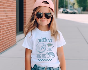 May include: A white t-shirt with a blue graphic of a rat and the text "THE RAT" and a list of personality traits. The model is wearing a pink baseball cap and brown sunglasses.