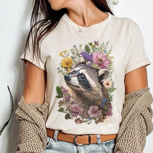 May include: A beige t-shirt with a graphic print of a raccoon surrounded by colorful flowers. The raccoon is looking up and has a black nose and eyes.