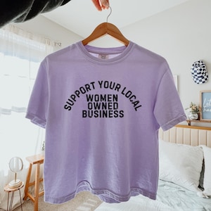 May include: A lavender t-shirt with the text "Support Your Local Women Owned Business" printed in black.