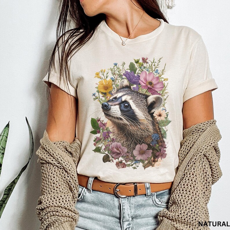 May include: A beige t-shirt with a graphic print of a raccoon surrounded by colorful flowers. The raccoon is looking up and has a black nose and eyes.