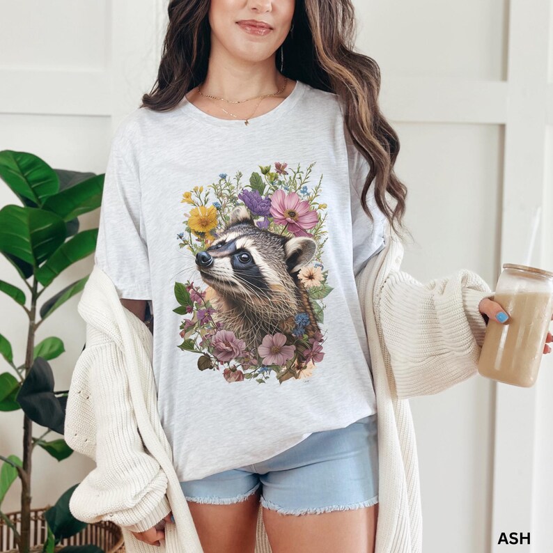 May include: A gray t-shirt with a floral design featuring a raccoon. The raccoon is surrounded by pink, purple, and yellow flowers.
