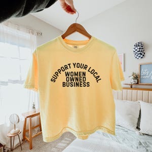 May include: A yellow cropped t-shirt with the text "Support Your Local Women Owned Business" printed in black.