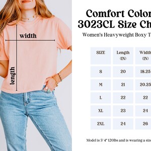 May include: Size chart for a women's heavyweight boxy t-shirt. The chart shows the length and width in inches for sizes S, M, L, XL, and 2XL. The model is 5'4" and wearing a size M.