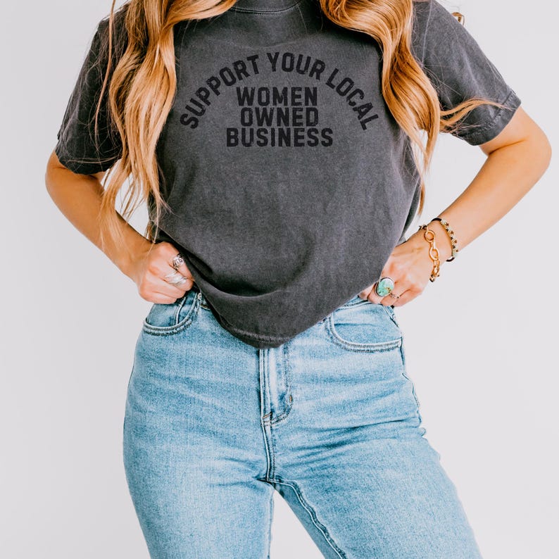 May include: A woman wearing a dark gray t-shirt with the text "Support Your Local Women Owned Business" printed on it. She is wearing blue jeans.