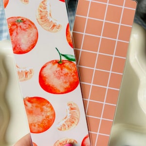May include: Two rectangular bookmarks. One features watercolor illustrations of oranges and orange slices on a white background. The other has a peach-colored grid pattern. The bookmarks are held in a hand.