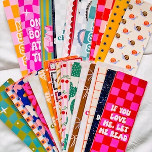 May include: A collection of colorful bookmarks with various designs. Some bookmarks feature text like "ON BOOKS AT TI" and "IF YOU LOVE ME, LET ME READ." Designs include checkered patterns, flowers, snails, and cacti. The bookmarks are arranged on a white surface.
