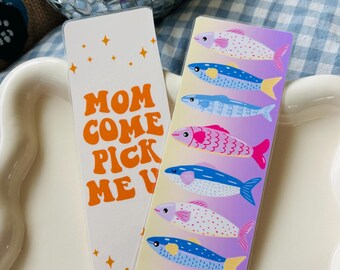 May include: Two rectangular bookmarks. One white bookmark has orange text that reads "MOM COME PICK ME UP" with orange star accents. The other bookmark has a gradient background with colorful fish illustrations.
