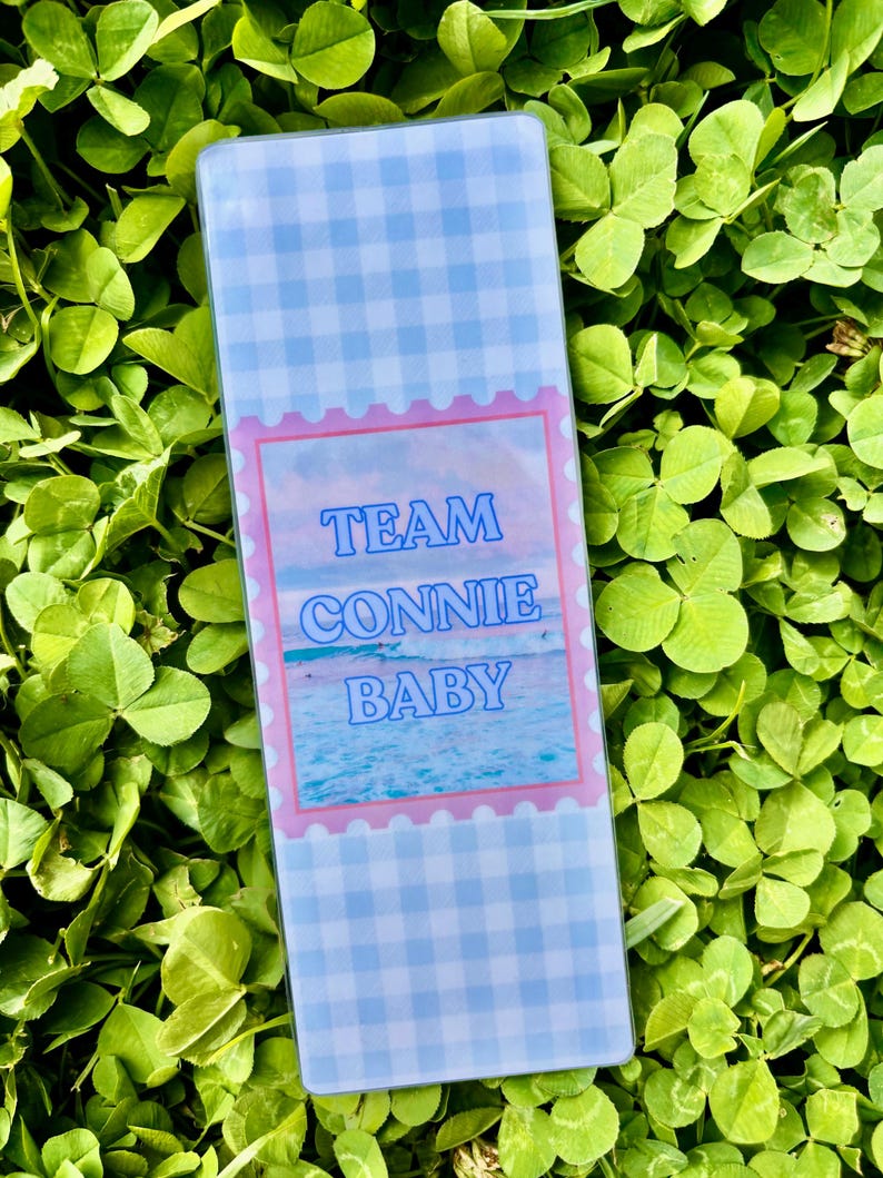 K&ouml;nnte beinhalten: Ein rechteckiger, klarer Acryl-Lesezeichen mit einem blau-wei&szlig;en Vichy-Muster. In der Mitte befindet sich ein rosa umrandetes Bild mit den Worten "TEAM CONNIE BABY" in Blau, vor einer Strandszene. Das Lesezeichen ist von gr&uuml;nem Laub umgeben.