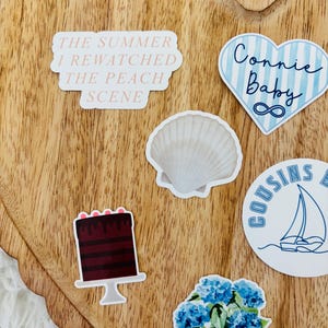May include: A collection of stickers on a wooden heart-shaped tray. Stickers include text that reads "The Summer I Rewatched The Peach Scene", a blue heart with "Connie Baby" and an infinity symbol, a seashell, a cake, a sailboat, and a potted plant.