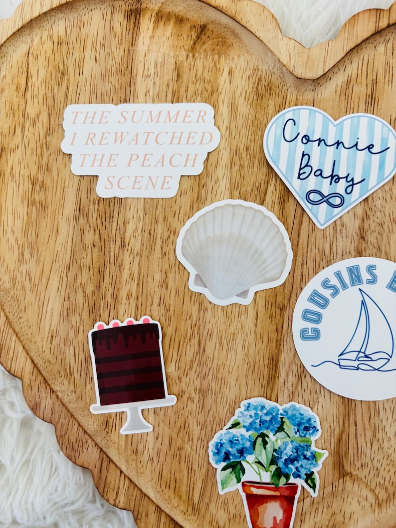 May include: A collection of stickers on a wooden heart-shaped tray. Stickers include text that reads "The Summer I Rewatched The Peach Scene", a blue heart with "Connie Baby" and an infinity symbol, a seashell, a cake, a sailboat, and a potted plant.