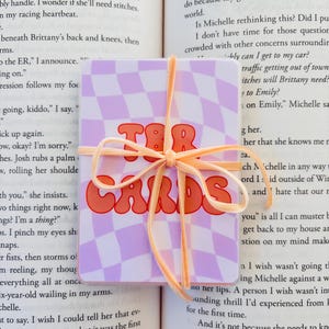 May include: A square card with the text "TBR CARDS" in orange letters, set against a lavender and white checkered background. The card is tied with a peach ribbon and rests on an open book.